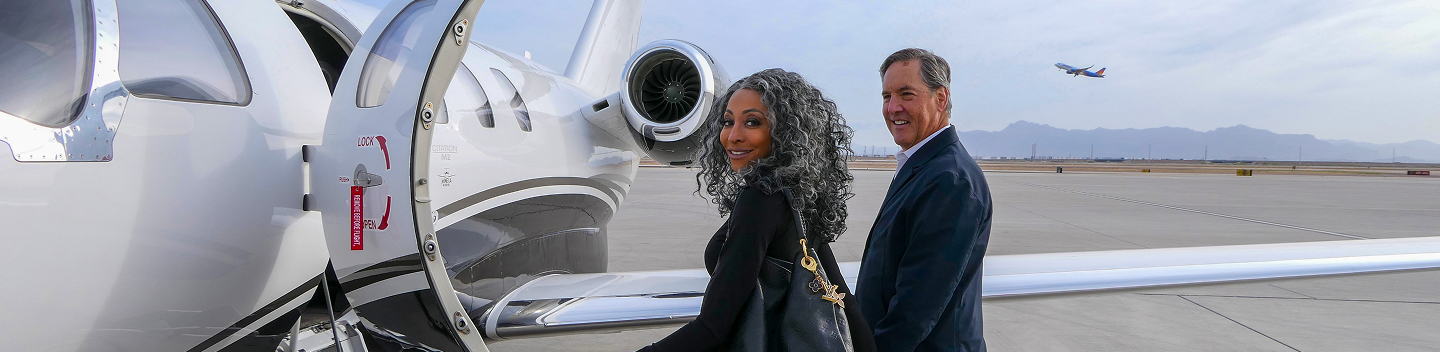 A smiling man and woman stand next to a white private jet with an open door on an airport tarmac. A commercial plane takes off in the distance.