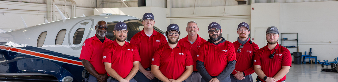 Group of eight men in red shirts and caps standing in front of a white aircraft in a hangar.