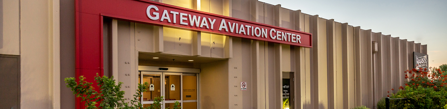 Exterior of the Gateway Aviation Center building with a large red sign and automatic glass doors. Green bushes and red flowers are in the foreground.