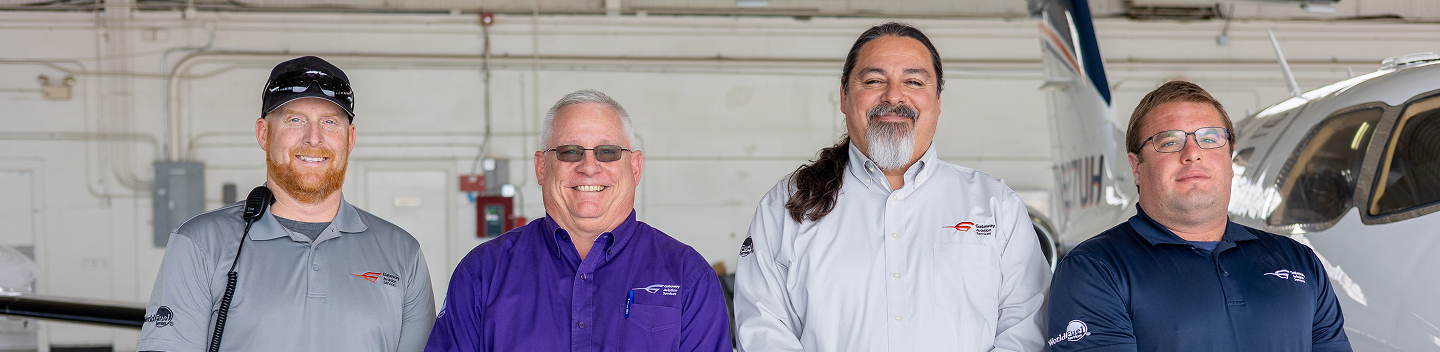 Four smiling men, dressed in branded shirts, stand in an aircraft hangar with parts of planes visible in the background.