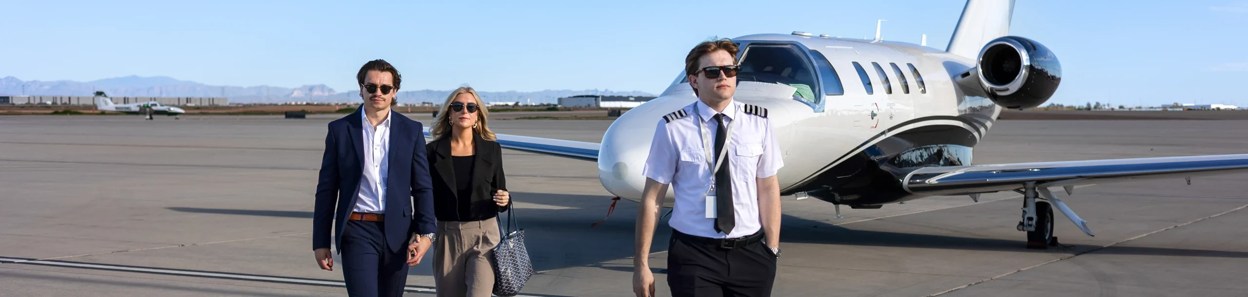 Three people, two passengers and a pilot, walk on an airport tarmac in front of a white and black private jet under a clear sky.